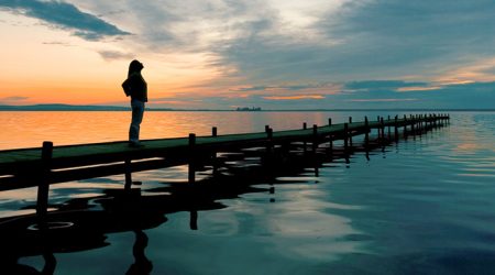 Young woman standing lakeside on jetty having a look at magical cloudscape and sunset colors.