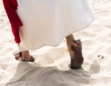 cropped image of Jesus in robe, sandals and red sash walking on sand in desert