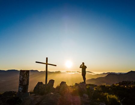 Male Silhouette Hiker standing on mountain summit next to a cross and cairn beacon taking a photo with a smart phone on a mountain summit at sunrise, Simonsberg, Stellenbosch, Cape Winelands, Western Cape, South Africa.